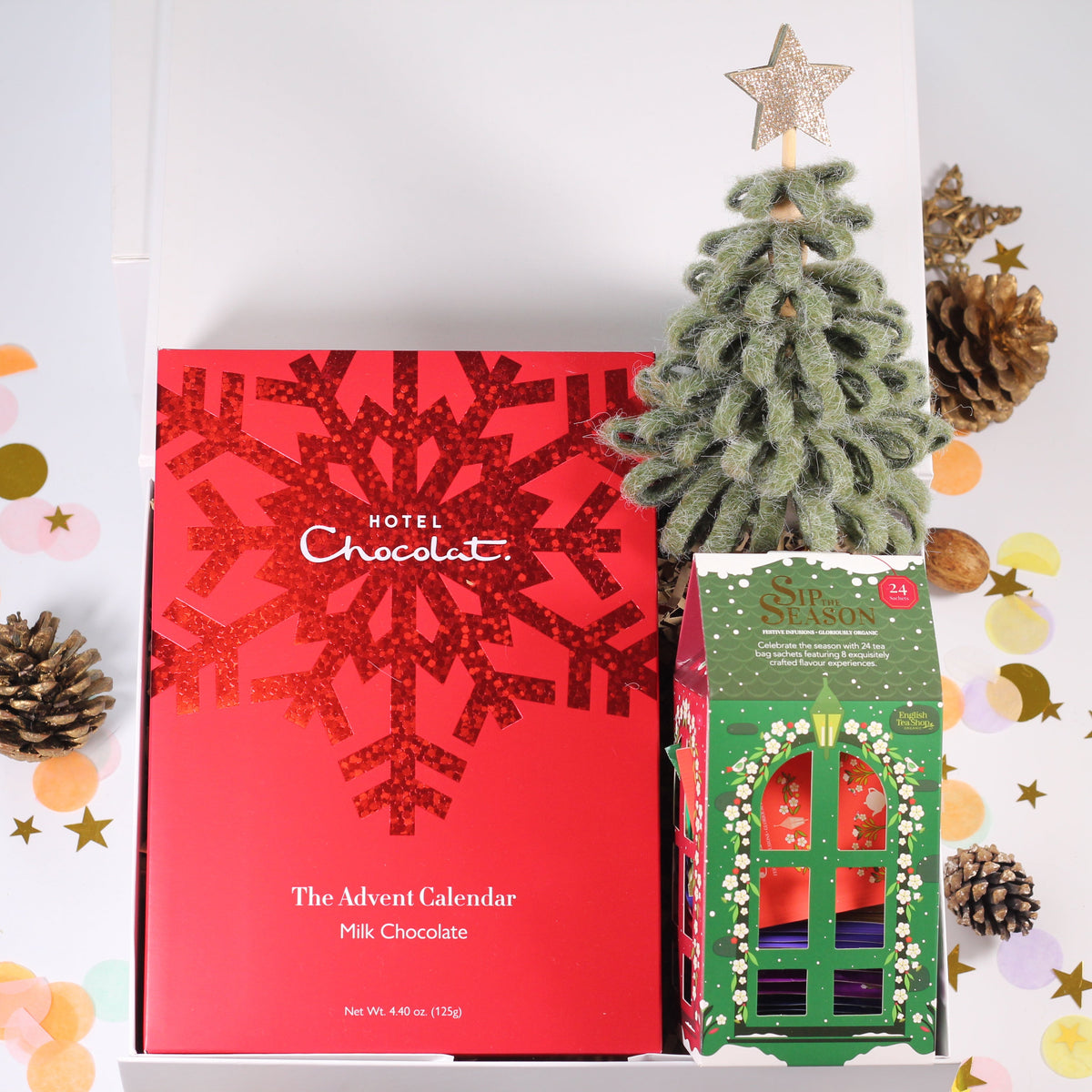 Hotel Chocolat The Advent Calendar Pink Chocolate box in red with a glittery snowflake pattern, displayed next to a small green Christmas tree and gift box inside an open white gift box, decorated with gold and pastel confetti and pinecones.