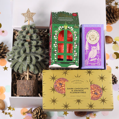 A curated gift box set from The Gift Shop of London, featuring a small artificial Christmas tree, a box of red chocolates in green packaging, a purple owl box, and a gold box of dark chocolate orange flowerpots, styled with pinecones and confetti on a white background.