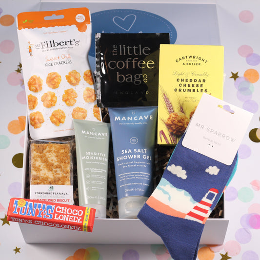 A variety of products including socks, shower gel, moisturizer, chocolate bar, biscuits, rice crackers, cheese crumbles, and coffee bags, neatly arranged in a keepsake magnetic box with a 'Box of Treats Just For Him' title.