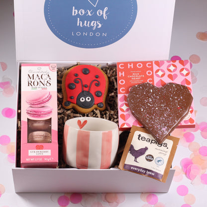 Gift box with macarons, ladybird cookie, a pink stripe mug, and a teabag and a heart chocolate on a colorful background.