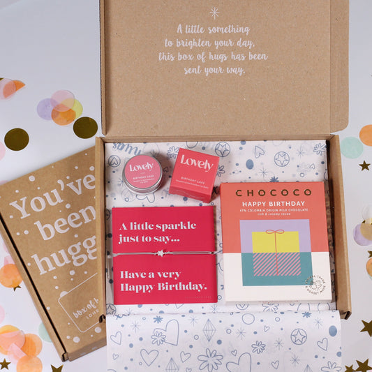 An open brown cardboard gift box on a white background with confetti and star decorations. Inside is a matching carded box containing a brown kraft card reading 'You've been hugged', a red birthday card, pink round items labeled 'Love' and 'Touch', and a chocolate box labeled 'CHOCOCO'.
