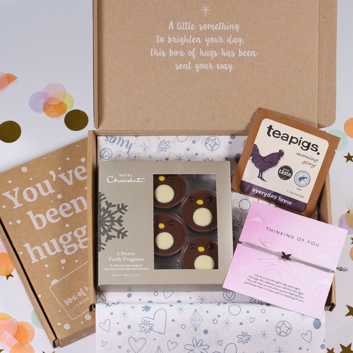 A curated gift box set displayed on a white surface with pastel confetti. The set includes a brown kraft box with a message, a book or card reading 'You've been hugged', a box of chocolate-covered treats, a small Teapigs tea box, and a pink handwritten note.