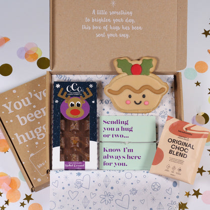 A curated gift box set displayed on a white surface with pastel confetti and star decorations. The set includes a kraft paper box with a printed message, a brown paper bag reading 'You've been hugged', a chocolate bar in a dark blue reindeer-themed wrapper, a small green card, a paper reindeer decoration, and a pouch labeled 'ORIGINAL CHOC CHOC BLEND'.