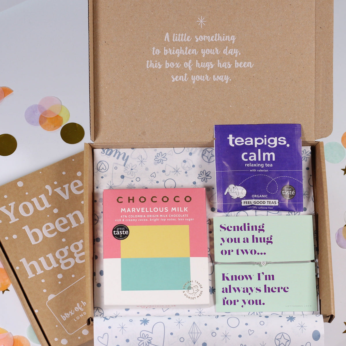 An open kraft gift box arranged on a white surface with scattered pastel confetti and gold star decorations. Inside the box are several small packaged items, including a purple tea bag packet labeled 'teapigs calm,' a pink and yellow card from 'CHOCOCO,' a mint green card reading 'Sending you a hug or two.