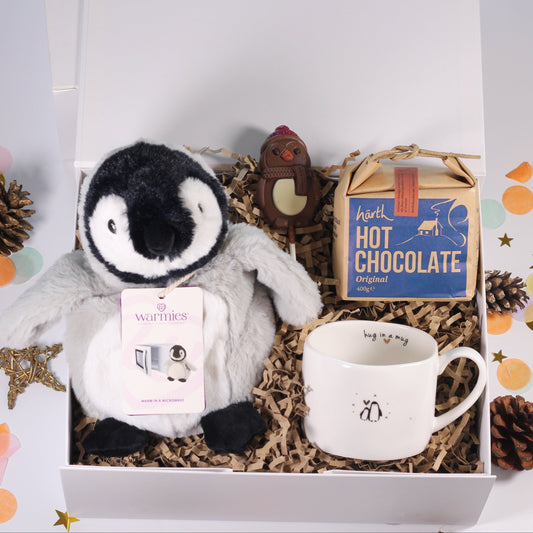 A curated gift box set featuring a plush penguin toy, a white ceramic mug with a penguin graphic, a box of chocolate hot chocolate, and a small wooden ornament, arranged in shredded paper inside an open white gift box with decorative confetti on a white background.