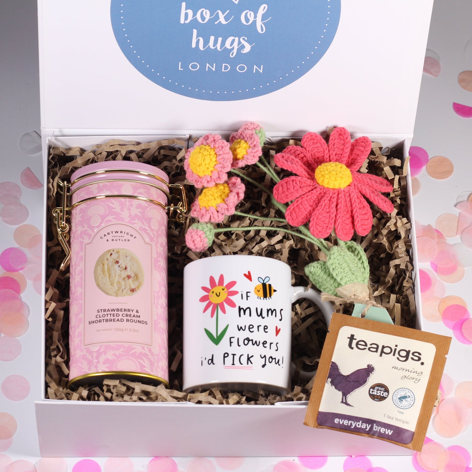 Gift set with a mum mug, tea sachet a tin of strawberry and clotted cream biscuits some pink crochet flowers in a box labeled 'box of hugs London'.