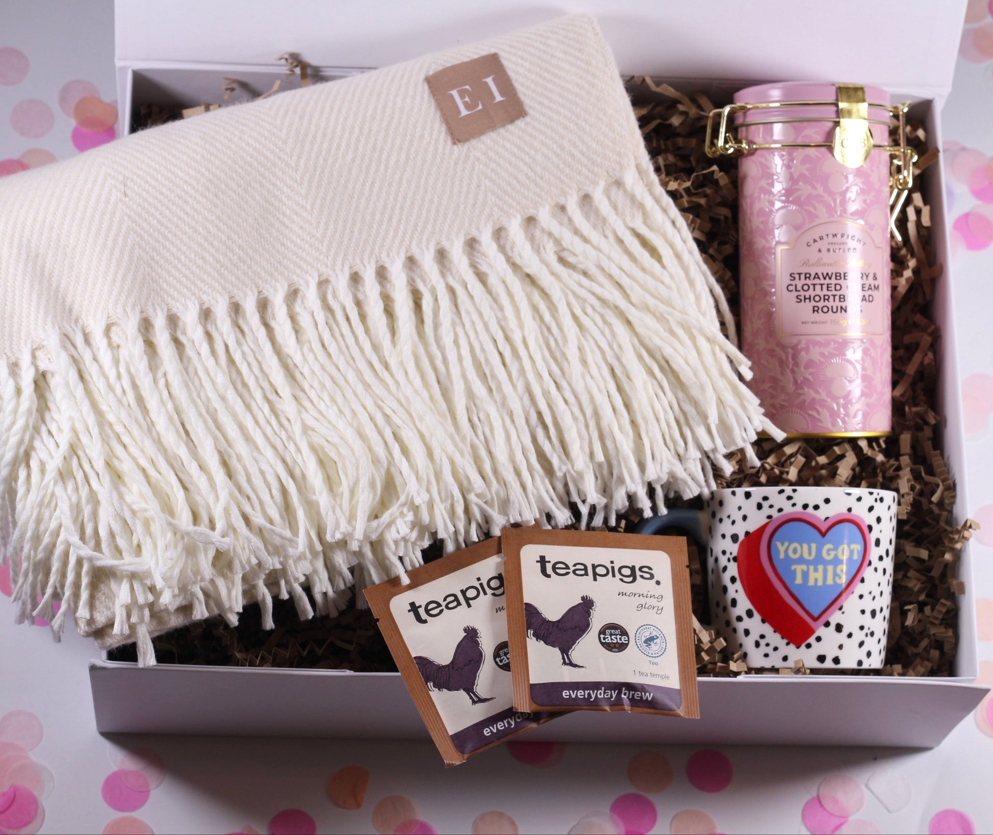 Gift box with a white blanket, teapigs tea bags, and strawberry and clotted cream shortbread and a you've got this mug, on a polka dot background