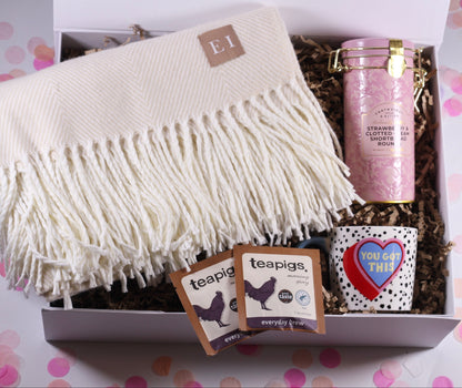 Gift box with a white blanket, teapigs tea bags, and strawberry and clotted cream shortbread and a you've got this mug, on a polka dot background