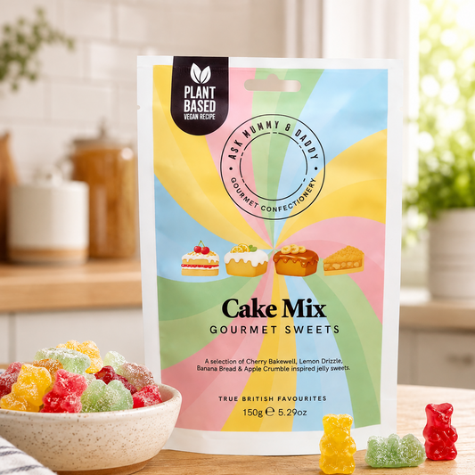 A stand-up pouch of Plant Based Vegan Foods Cake Mix Gourmet Sweets stands on a wooden table in a bright kitchen setting. A bowl of colorful gummy sweets sits to the left, and a few loose gummies are placed in front of the package.
