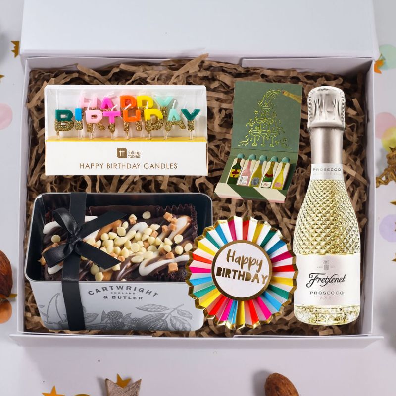 A curated birthday gift box set containing a bottle of Prosecco, a tin of gourmet chocolates, a box of birthday candles, a green card, and a colorful round 'Happy Birthday' badge, all arranged in brown paper shred inside a white gift box.
