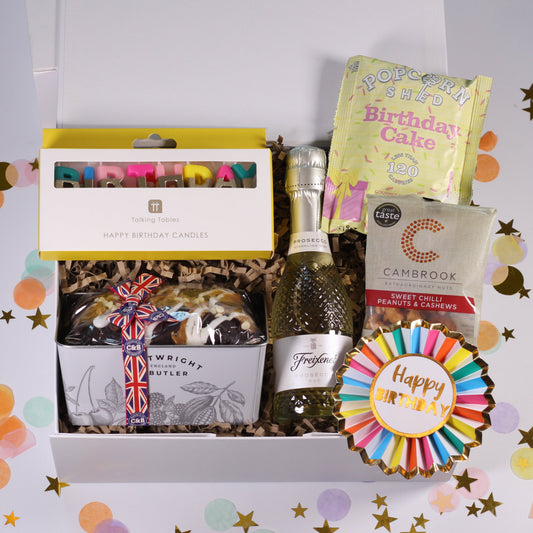 A curated birthday gift box filled with assorted treats, including a boxed candle, a bottle of sparkling wine, a cake, a bag of Cambridge tea, and a colorful 'Happy Birthday' fan ribbon, arranged on a white background with confetti.