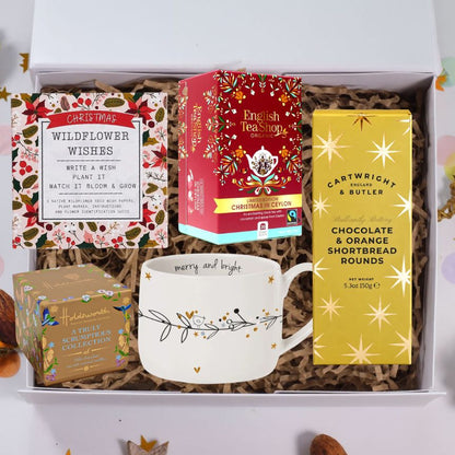 A curated gift box set displayed inside an open white box filled with brown paper shred. Visible items include a Christmas-themed wildflower wish card, a box of English Tea Shop Christmas Oolong tea, a yellow box of Cartwright & Butler chocolate and orange shortbread rounds, a small boxed collection of Hallmark winter scarves, and a white ceramic mug with gold and black botanical details.