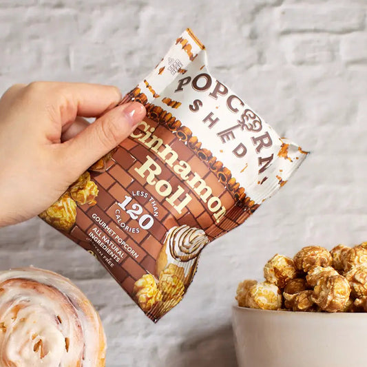 A hand holds a bag of Popcorn Shred Cinnamon Roll flavored popcorn in front of a white brick wall, with a cinnamon roll, a bowl of popcorn, and scattered popcorn kernels on a marble surface.
