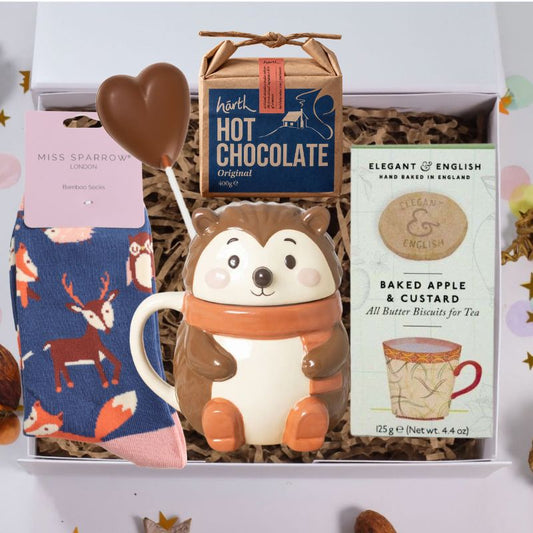 A curated gift box set containing a ceramic hedgehog mug with an orange scarf, a chocolate heart on a stick, a box of Hartley's Hot Chocolate, and a pair of blue patterned socks with deer and fox motifs.