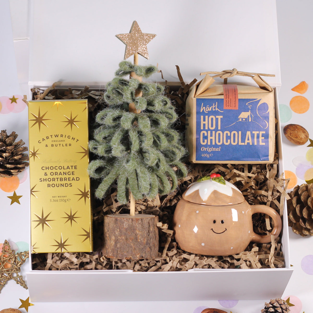 A curated gift box set featuring a small artificial Christmas tree, a box of Lavender & Butter chocolate shortbread, a bag of Mott Hot Chocolate, and a ceramic mug shaped like a gingerbread cookie, arranged in shredded paper inside an open white box with decorative confetti and pinecones around it.