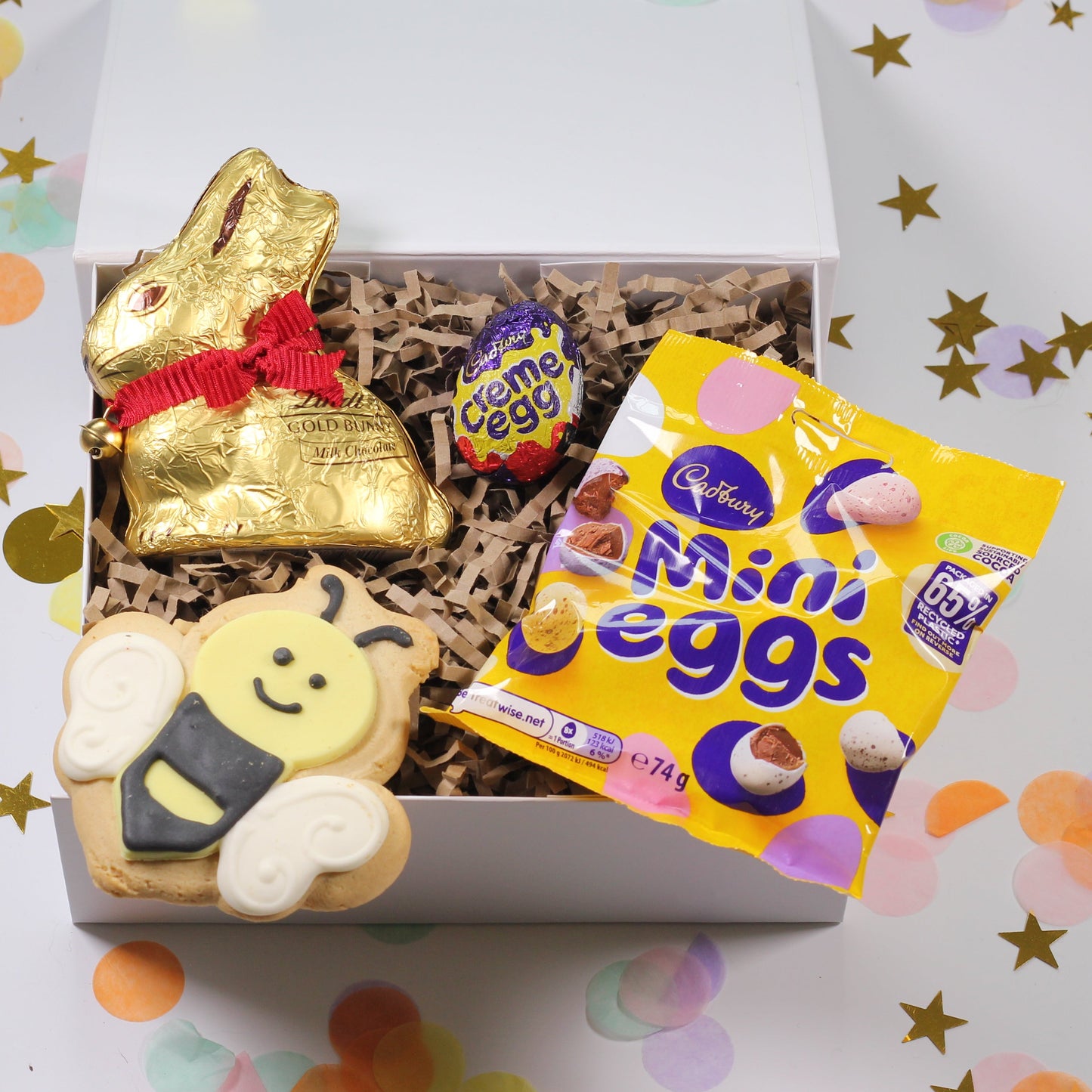 Gift box with Easter-themed items including a gold bunny, a bee-shaped cookie, and a bag of Cadbury mini eggs on a confetti background.
