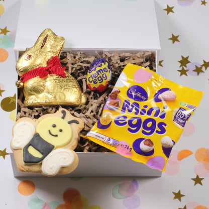 Gift box with Easter-themed items including a gold bunny, a bee-shaped cookie, and a bag of Cadbury mini eggs on a confetti background.