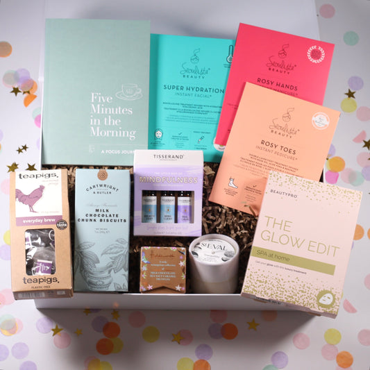 A collection of self-care products including a focus journal, roller ball essential oils, a pot candle, and other items, neatly arranged in an open white magnetic box with colorful dots pattern.