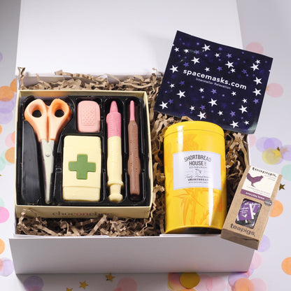 A gift box with various items including chocolate in the shape of medical supplies, a tin of shortbread biscuits, a self-heating eye mask, and a tea package, all presented in a white magnetic box with a get well card.