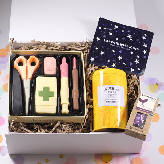 A gift box with various items including chocolate in the shape of medical supplies, a tin of shortbread biscuits, a self-heating eye mask, and a tea package, all presented in a white magnetic box with a get well card.