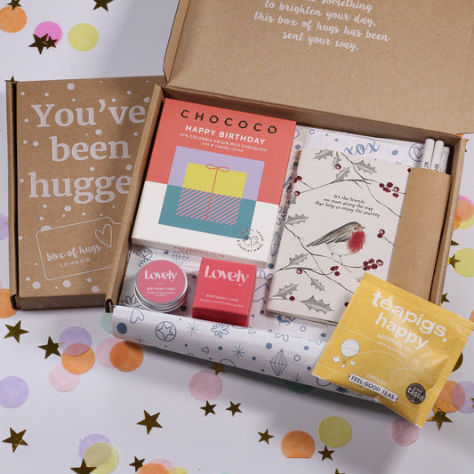 A letterbox gift set containing a variety of items including a card, a notebook, pencils, a chocolate bar, lip balm, and a tea sachet, presented in a box with a 'You've Been Hugged' design.