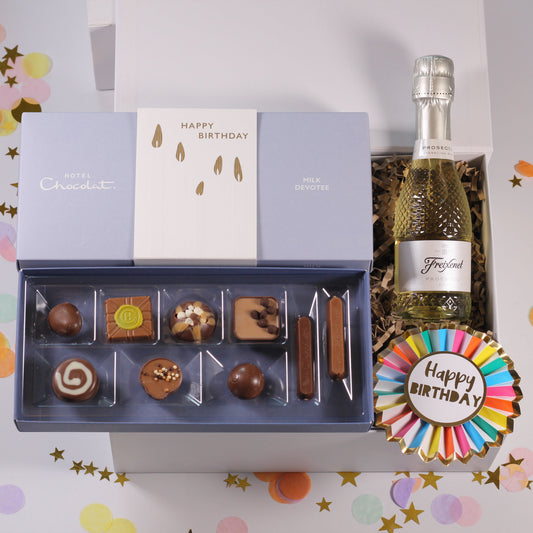 A luxury chocolate and champagne gift set arranged in an open white box on a confetti-covered surface. The set includes an open blue-lavender chocolate tray with assorted chocolates, a bottle of Trifield sparkling wine, and a round multicolor 'Happy Birthday' ribbon decoration.
