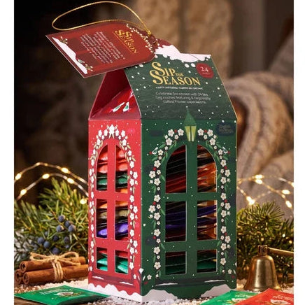A Christmas-themed gift box set displayed on a wooden surface in a cozy indoor setting. The main product is a house-shaped box labeled 'Spiced Season' with red and green sides, white snowflake details, and colorful contents visible through cutout windows.