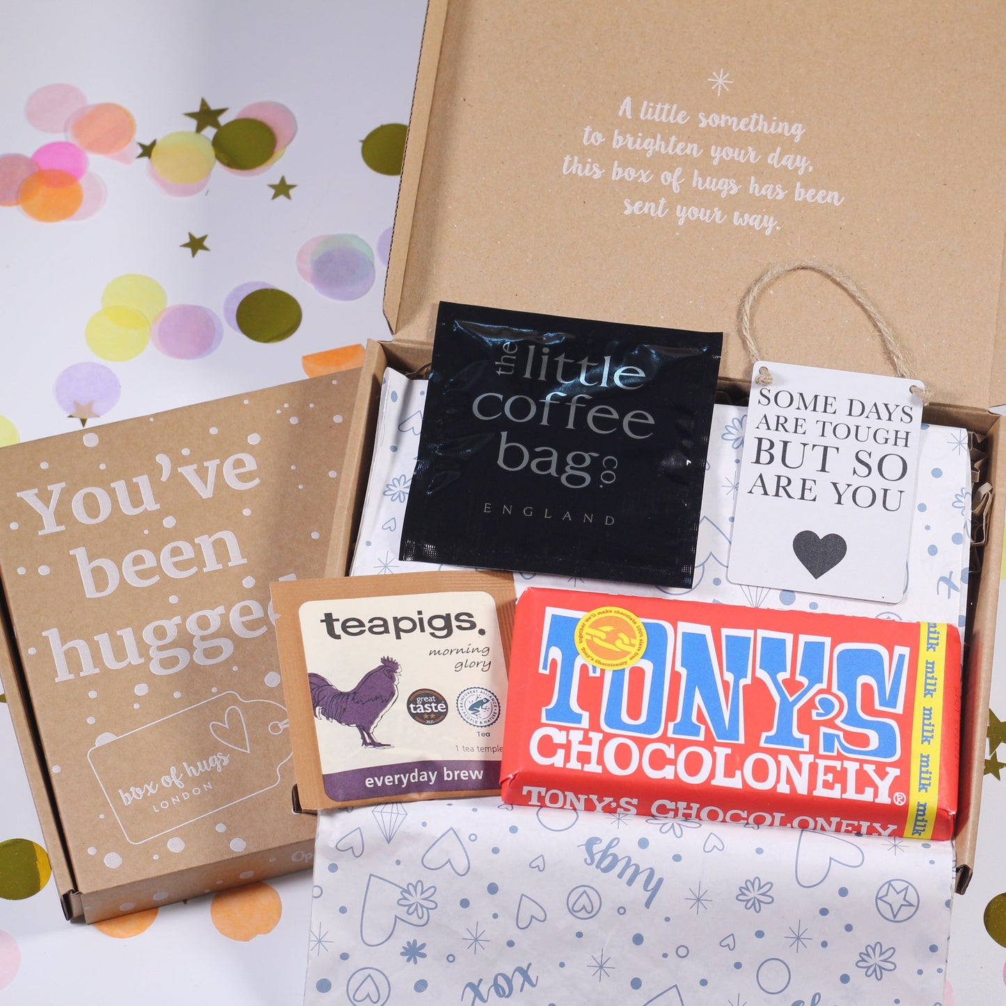 A letterbox gift hamper containing a variety of items including a chocolate bar, tea bags, coffee bags, a motivational mini sign, and an eye mask, all packaged in a brown box with a 'You've Been Hugged' note.