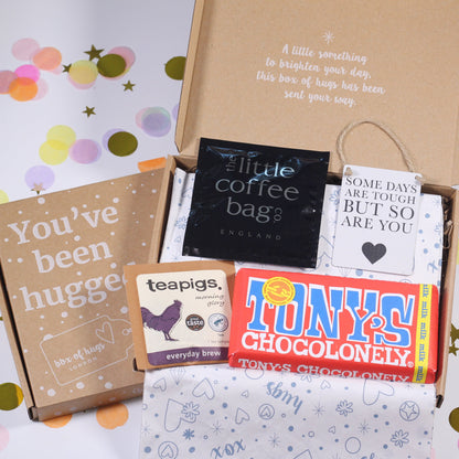 A letterbox gift hamper containing a variety of items including a chocolate bar, tea bags, coffee bags, a motivational mini sign, and an eye mask, all packaged in a brown box with a 'You've Been Hugged' note.