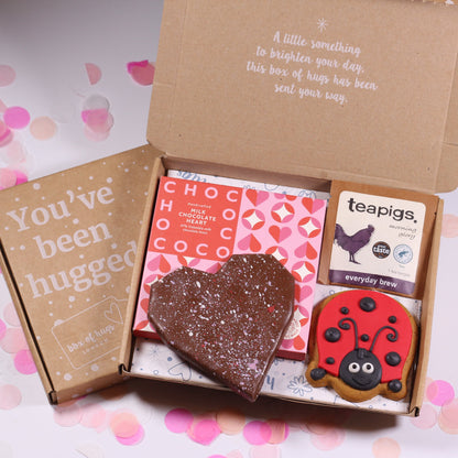 Open gift box with heart-shaped chocolate, teapigs tea, ladybird gingerbread biscuits, in a  'You've been hugged' letterbox on a pink and white background.