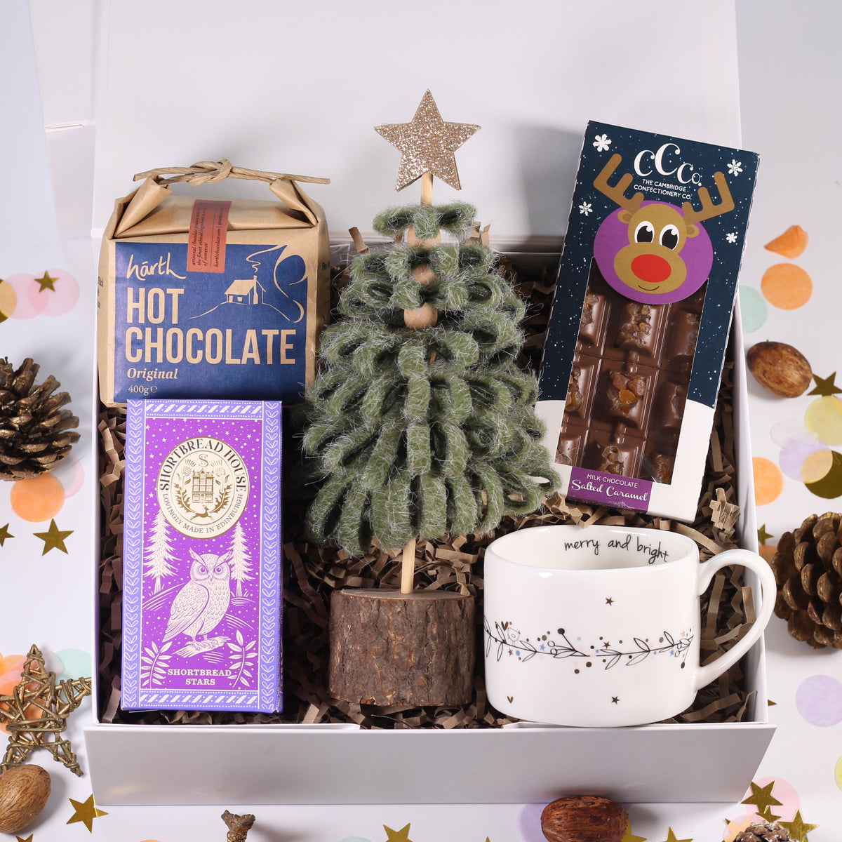 A curated gift box set on a white background with confetti and pinecones. The box contains a small fuzzy green Christmas tree, a purple owl hot chocolate box, a blue hot chocolate box, a white mug with a holiday design, and a packaged chocolate bar with a cartoon face.
