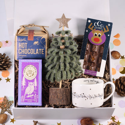 A curated gift box set on a white background with confetti and pinecones. The box contains a small fuzzy green Christmas tree, a purple owl hot chocolate box, a blue hot chocolate box, a white mug with a holiday design, and a packaged chocolate bar with a cartoon face.