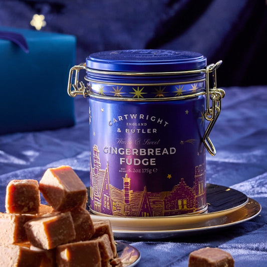 A tin of Cotwicky Heritage & Cutler Gingerbread Fudge sits on a saucer beside plates of cut fudge pieces, with a wrapped gift box blurred in the background on a blue quilted surface.