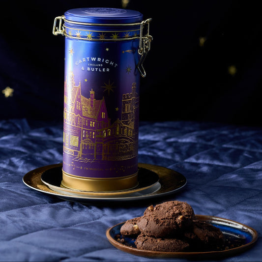 A cylindrical blue and purple tin with gold castle artwork and the text 'CASTLEBRIGHT BAKERY & SUGAR' stands on a small gold tray. A plate of chocolate cookies sits in front on a blue fabric surface against a dark blue starry backdrop.