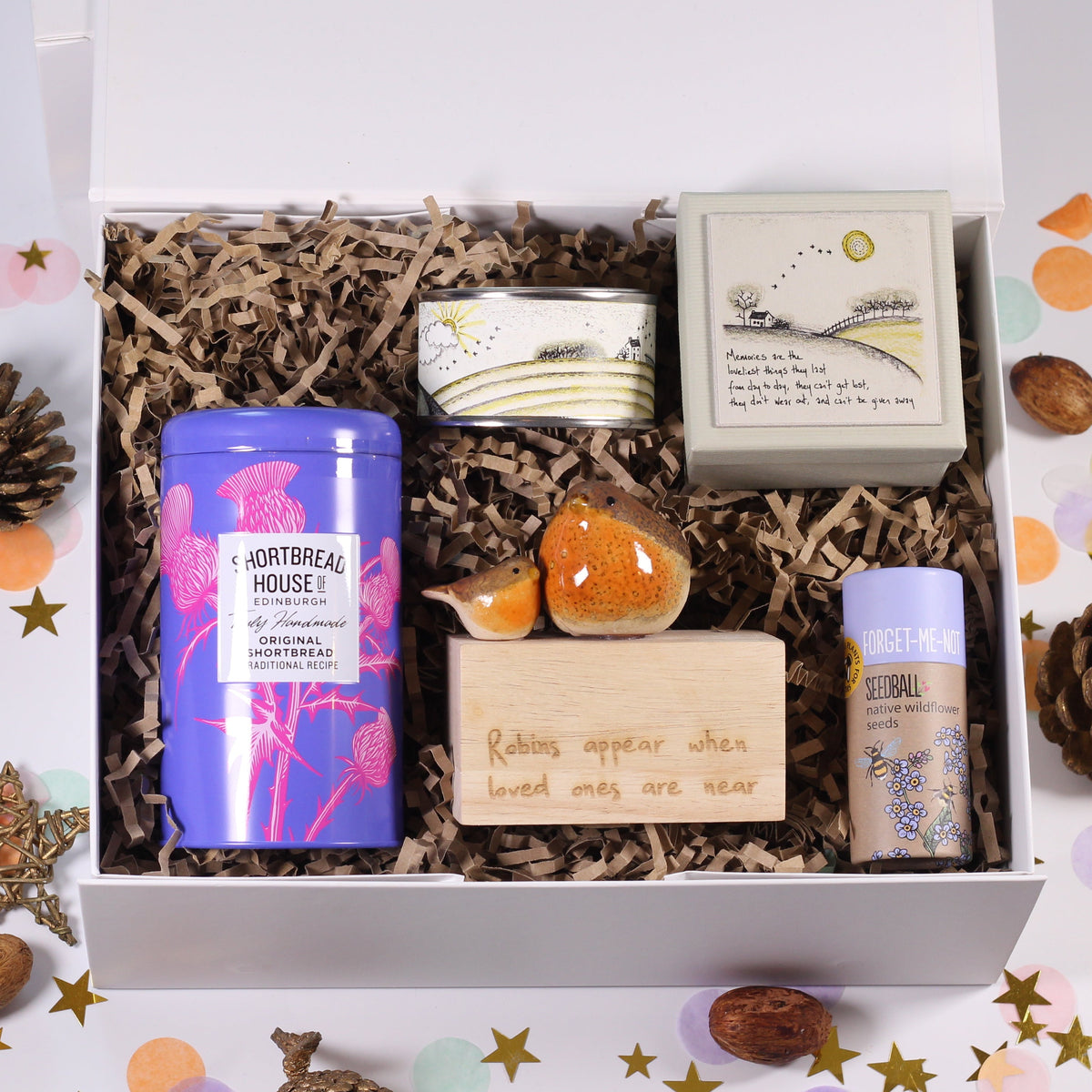 An open white gift box filled with natural housekeeping products, including a blue tin labeled 'Natural Housekeeping', a small wooden block, a candle, and a boxed item, arranged on a white background with scattered confetti and pinecones.