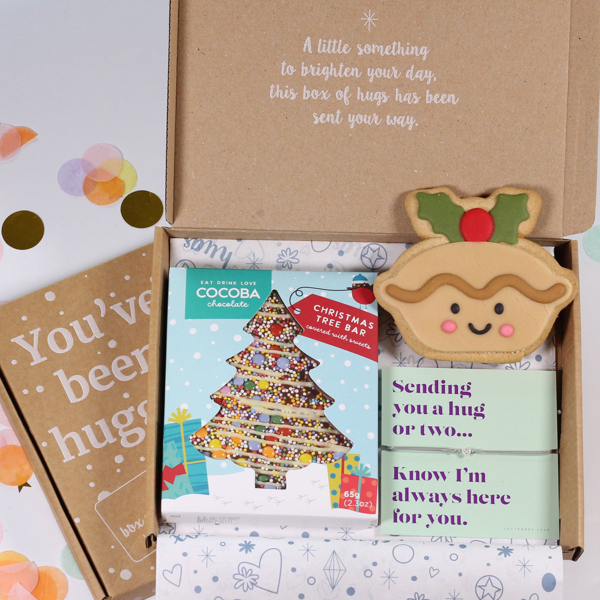 A Christmas-themed gift box set on a white background with gold and pastel confetti. The open kraft cardboard box contains a wrapped chocolate bar labeled 'COCOA', a green card reading 'sending you a hug or two.
