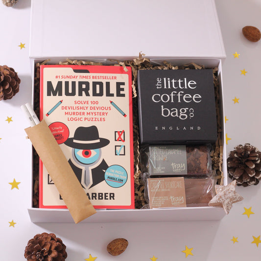 A white magnetic box with 'MURDLE' puzzle book, a chocolate bar, a brownie, and a set of pencils displayed inside. The box is accompanied by a 'The Little Coffee Bag' black card and pine cones scattered around.