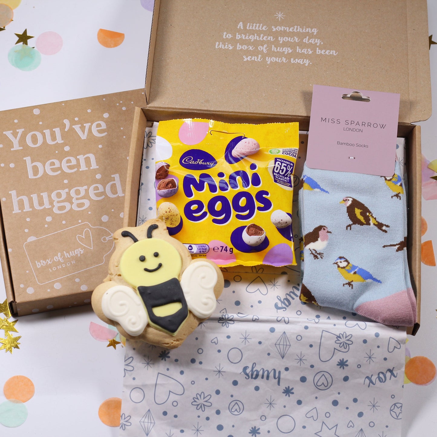 Gift box with Easter-themed items including Mini Eggs, an iced bee biscuit, some bird print socks on a white background with confetti.