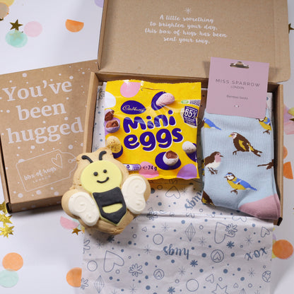 Gift box with Easter-themed items including Mini Eggs, an iced bee biscuit, some bird print socks on a white background with confetti.