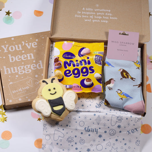 Gift box with Easter-themed items including Mini Eggs, an iced bee biscuit, some bird print socks on a white background with confetti.