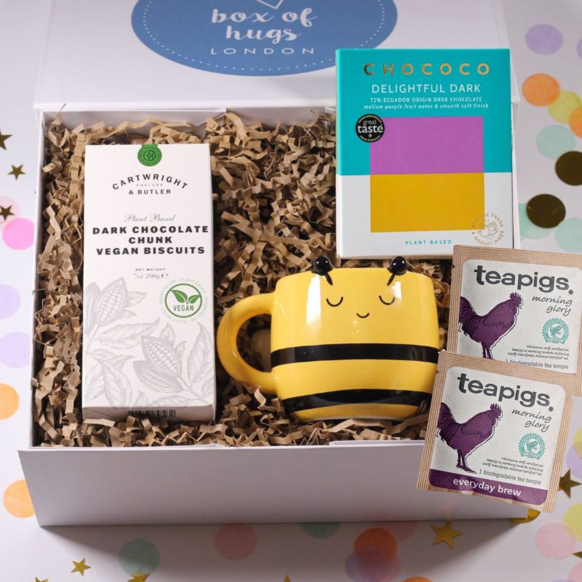Gift box with a bee-shaped mug, dark chocolate bar, and teapigs and vegan biscuits on a colorful background.
