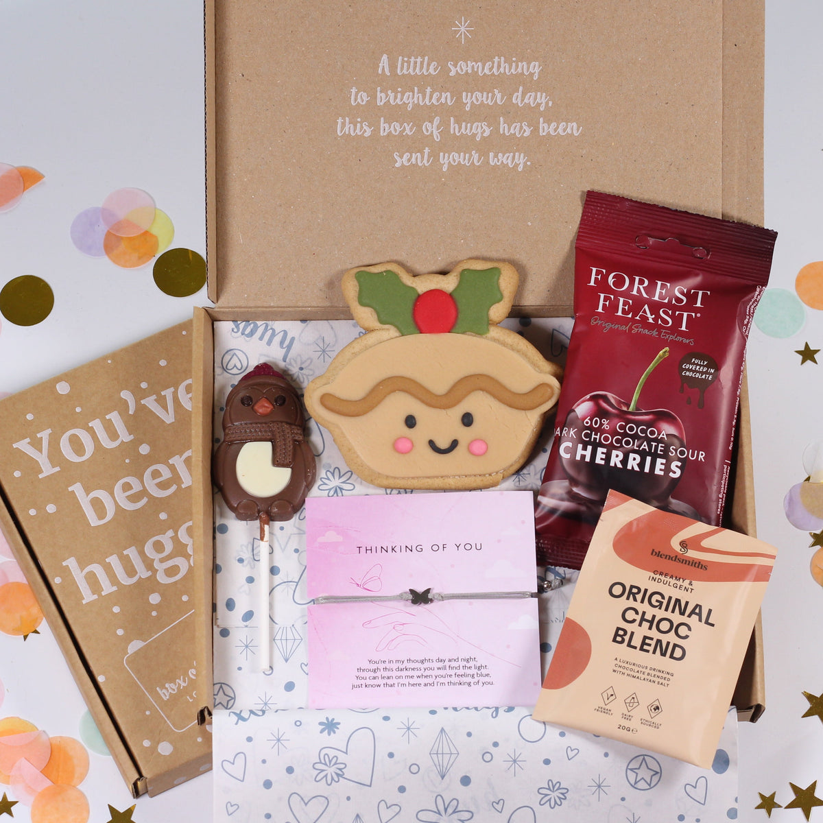 A curated gift box set displayed in an open kraft cardboard box on a white surface with pastel confetti. Visible items include a brown paper book or journal with the phrase 'You've been hugger', a small chocolate-covered stick treat, a pink card, a round decorated cookie, a red Forest Feast chocolate chip cookie bag, and a bag of Original Choc Choc Blend.