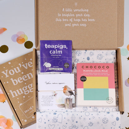 An open kraft gift box arranged on a white surface with pastel confetti and gold star decorations. Inside the box are several small items: a purple Teapigs tea box, a pink and yellow Chococo Flavours box, a brown card that says 'You've been hugged', and a patterned tissue box.