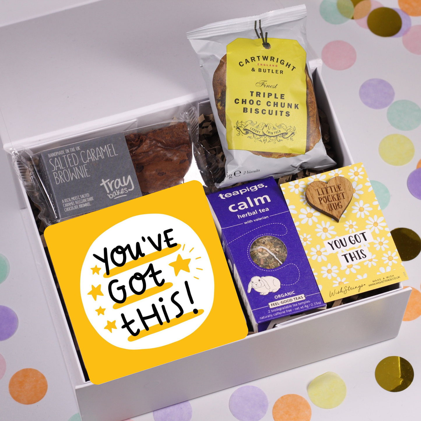 A gift box of various items including a chocolate brownie, biscuits, tea temples, a motivational coaster, and a keepsake card with a message, all arranged in a white box with confetti background.