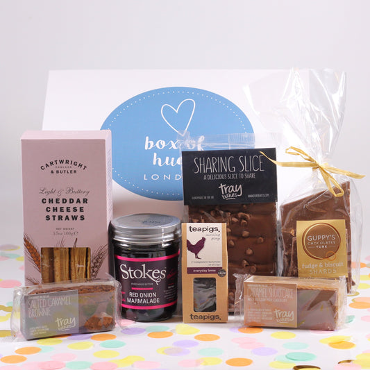 An assortment of food items including chocolate, biscuits, brownies, and a cheese straws, neatly arranged on a colorful surface, all packaged in a white magnetic box with a blue and red heart design on the lid, accompanied by a selection of cards.