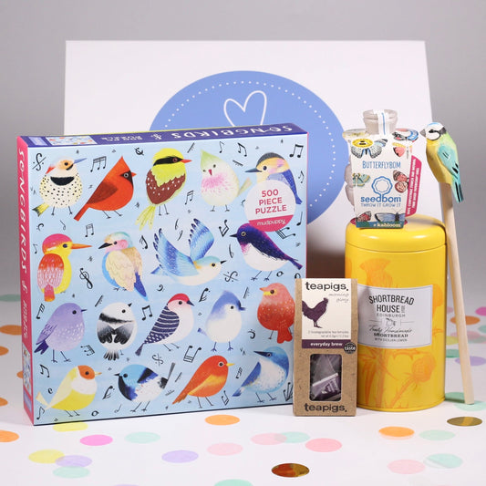 A variety of items including a bird-themed puzzle, a box of shortbread biscuits, a packet of pollinator bee seeds, a pencil with bird design, and tea bags, all arranged for photography.