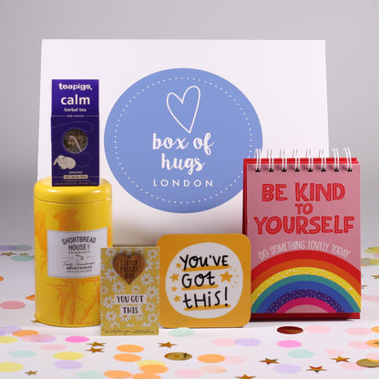 A gift box with a pocket hug keepsake token, a motivational coaster, a tin of lemon shortbread biscuits, and a self-care quote book.